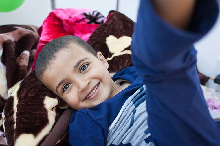 Young Hamam lays on his hospital bed, waiting for his lifesaving heart surgery. He has a big smile and his eyes sparkle.