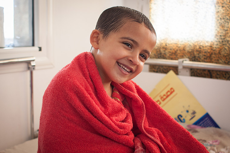 Young Hamam is wrapped up in a red towel, smiling. He just had a bath before his lifesaving heart surgery.