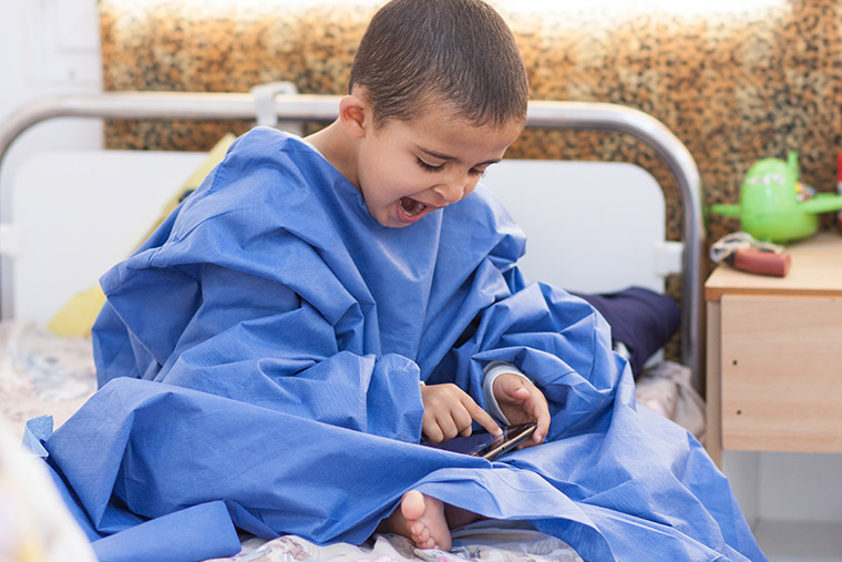 Hamam sits on his hospital bed, wrapped in a sterile gown, and yawning while playing a video game. Waiting for heart surgery is hard work!