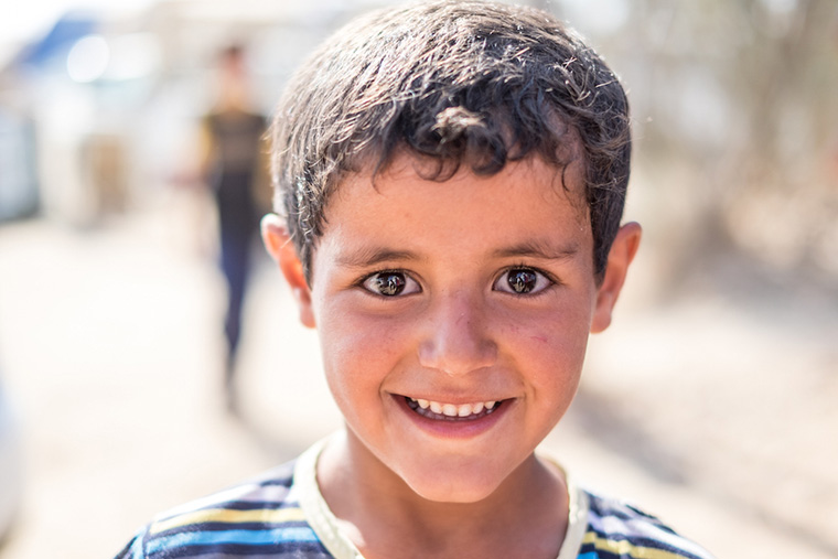 A young, bright-eyed boy poses with a smile. His family are displaced, camped out in the Iraqi desert, and in need of aid.