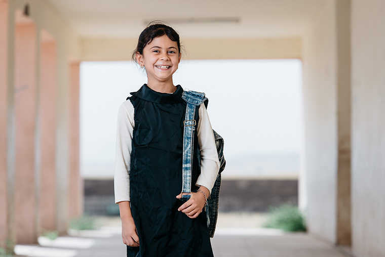 A young Iraqi girl poses in a hallway at her school. She is dressed in a uniform, backpack, and a big smile.