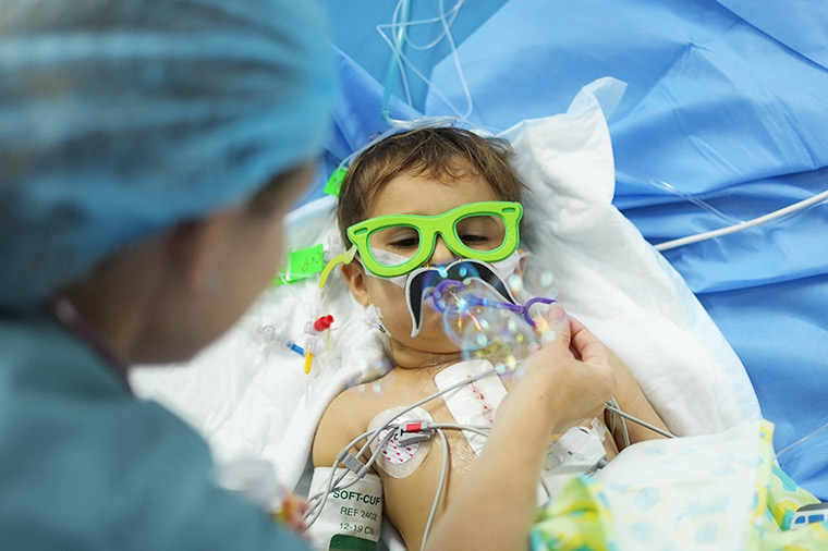 A nurse helps a young patient blow bubbles. He lays in his hospital bed, wired to machines, yet wearing large green foam glasses, just for fun.