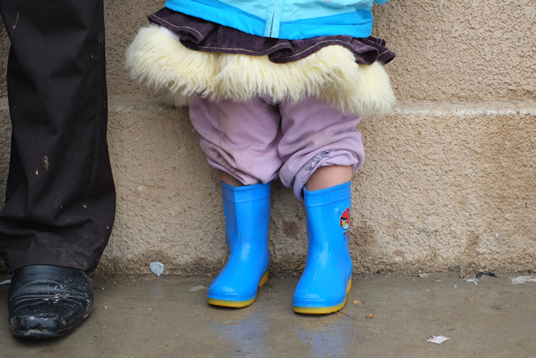 A young IDP girl stands next to her father on a cold rainy day. She wears the new, rain-proof boots we delivered to her community.