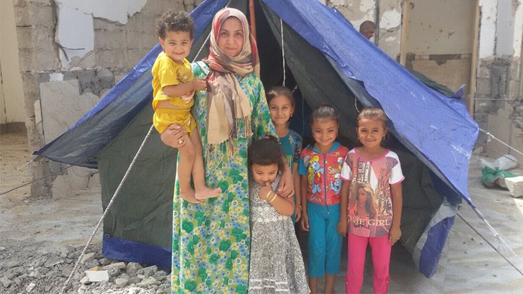 A mom and her young children stand at the entrance of their new tent. The mother smiles—she now has shelter for her children.